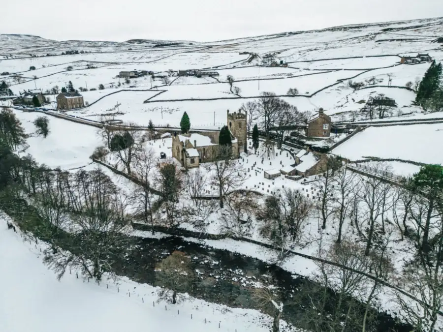 Snow over walled farmland, a church and a river in the north Pennine foothills, the winter weather that lies longest on roofs east of Carlisle