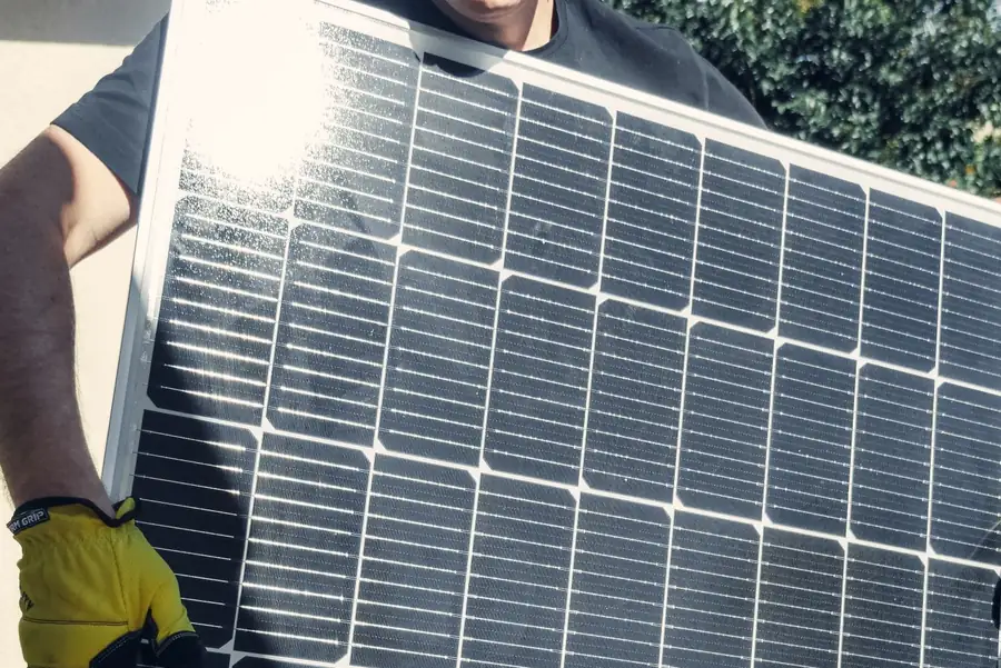 An installer carrying a single photovoltaic module by hand, the 20 to 25kg unit whose weight multiplies across a Carlisle roof slope