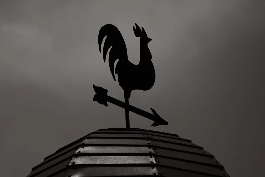 A weather vane on a ridge against heavy cloud, showing the prevailing wind that drives rain under the laps at the edges of a Carlisle roof